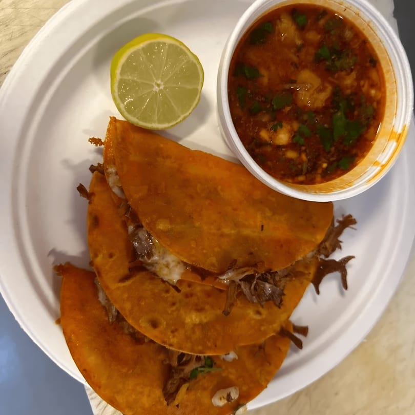 Three birria tacos with a side of consommé and a lime wedge on a white plate.